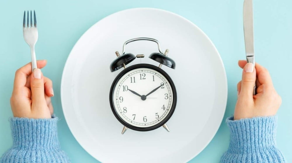 2 hands holding a knife and a fork combining with a white plate and a clock on top | Feature | 10 Reasons Why Intermittent Fasting Is The Simplest Diet Ever